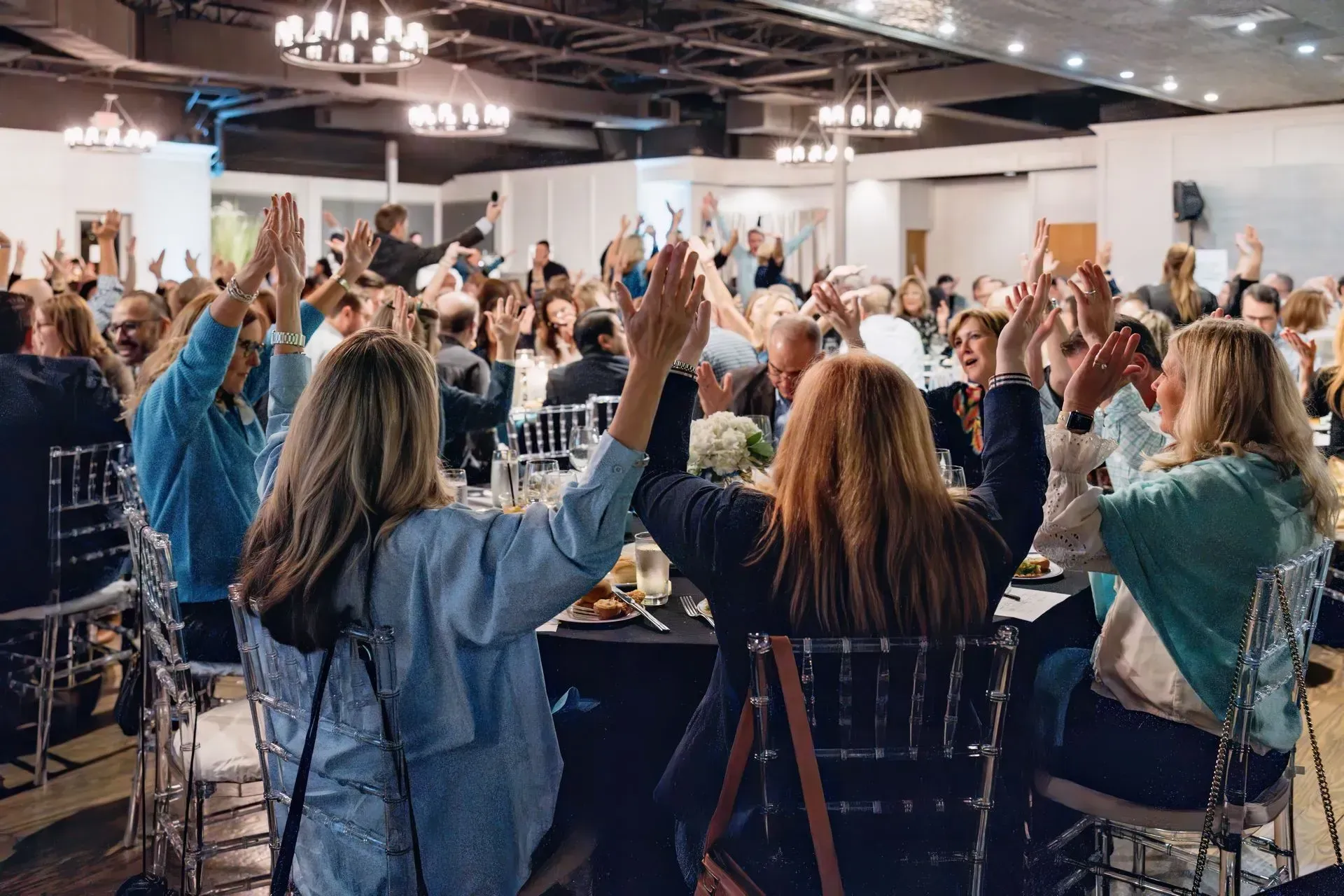 Conference attendees raising hands in a banquet hall with round tables and chandeliers