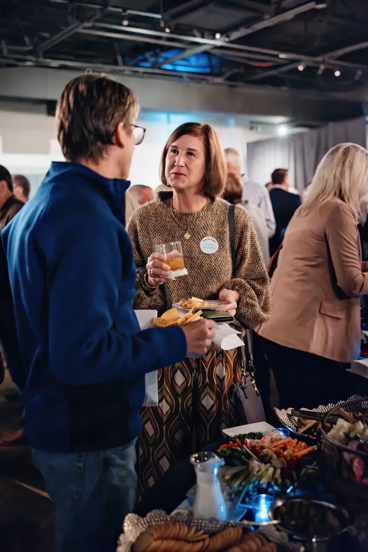 Two people chatting at a buffet table with plates of food in a bright indoor event space