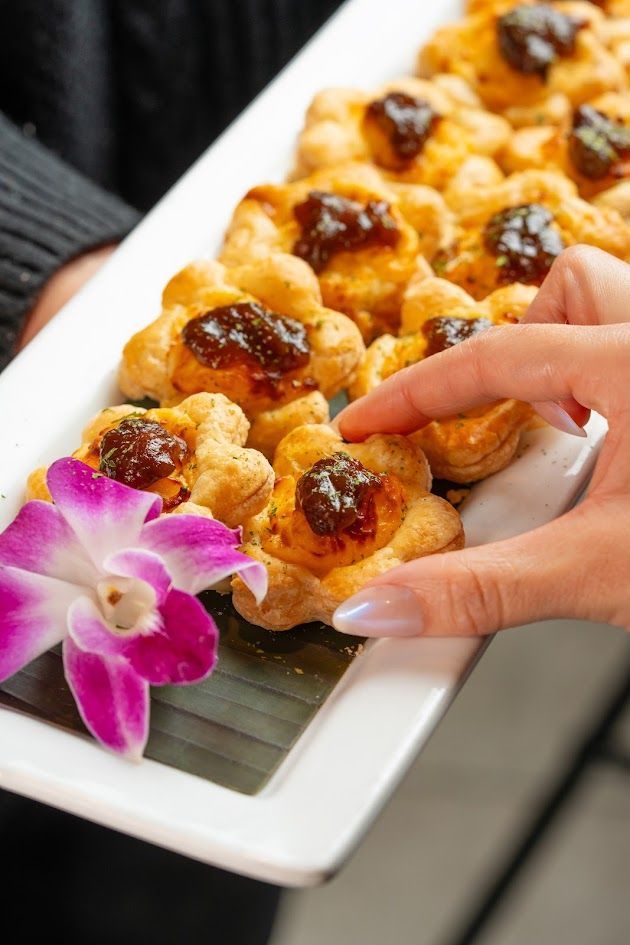 Hand placing a pastry on a white platter of glazed pastries with a purple orchid garnish