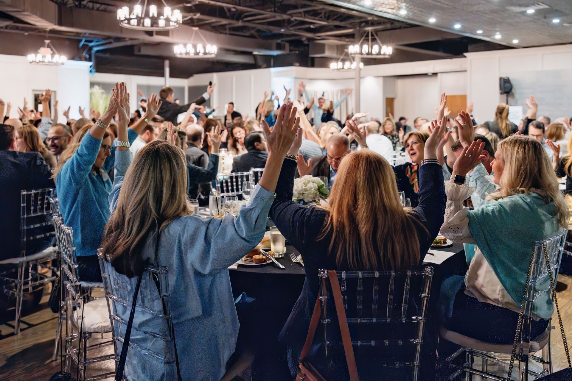 A large group of people are sitting at a table with their hands in the air.