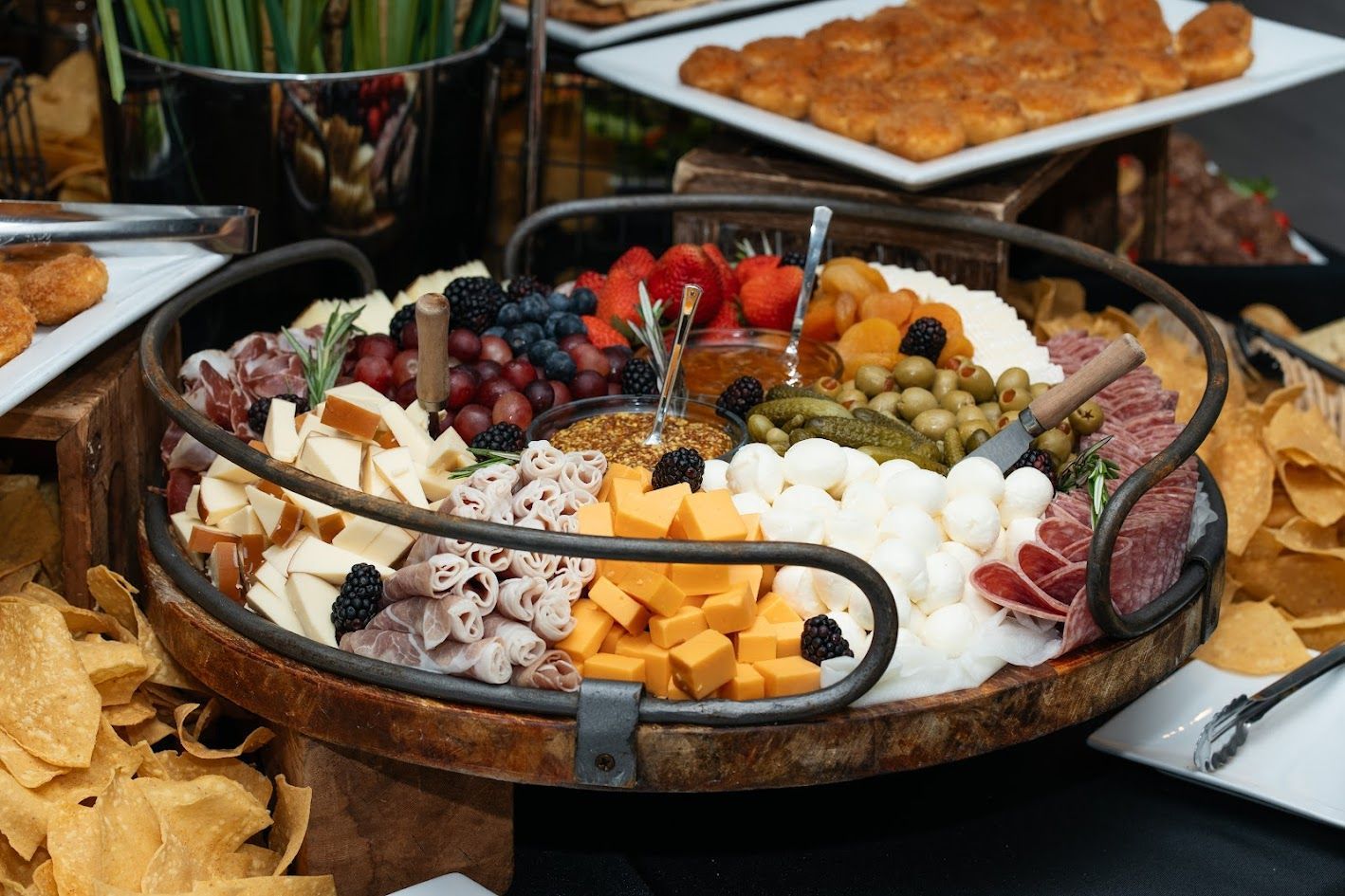 A wooden tray filled with meats, cheeses, vegetables, and chips on a table.