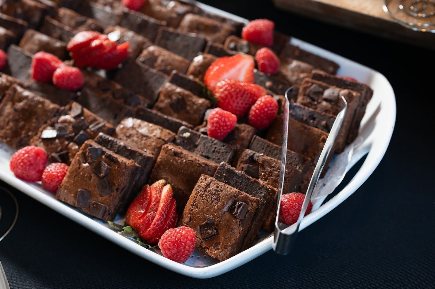 A white plate topped with brownies and raspberries with tongs.