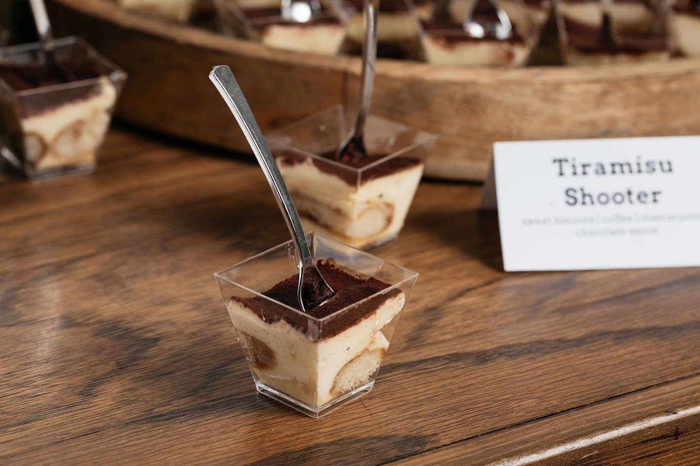 A tiramisu shooter with a spoon in it is on a wooden table.