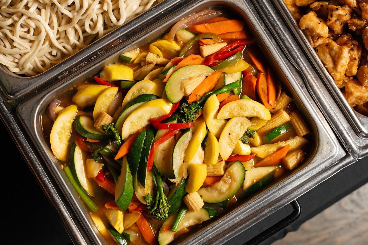 Colorful stir-fried vegetables in a black tray, with noodles and grilled meat on the side
