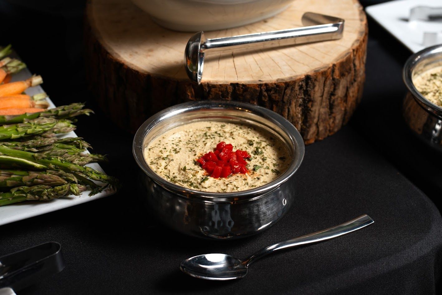 Metal bowl of creamy dip topped with red garnish, beside asparagus and spoon on a dark table