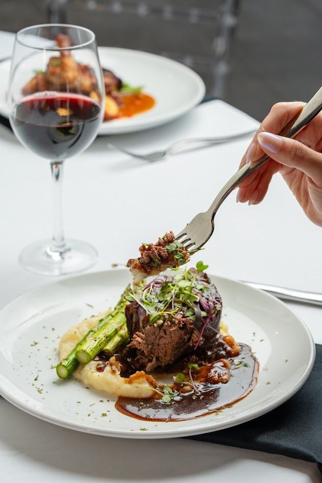 Hand placing a forkful of gourmet steak with asparagus and sauce on a white plate beside a glass of red wine