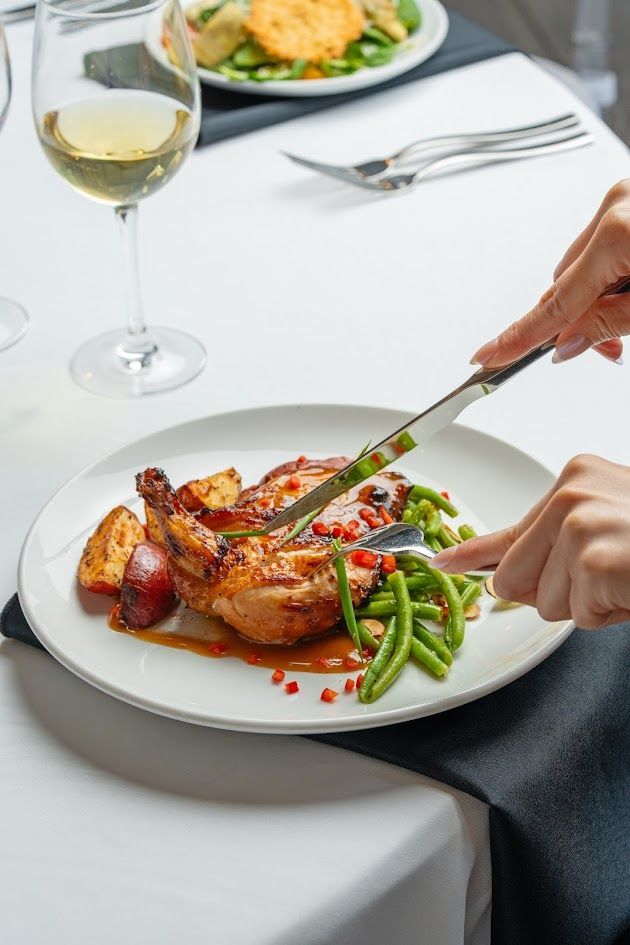 Dining table with plated chicken, vegetables, and wine, while hands cut the meal with a knife and fork