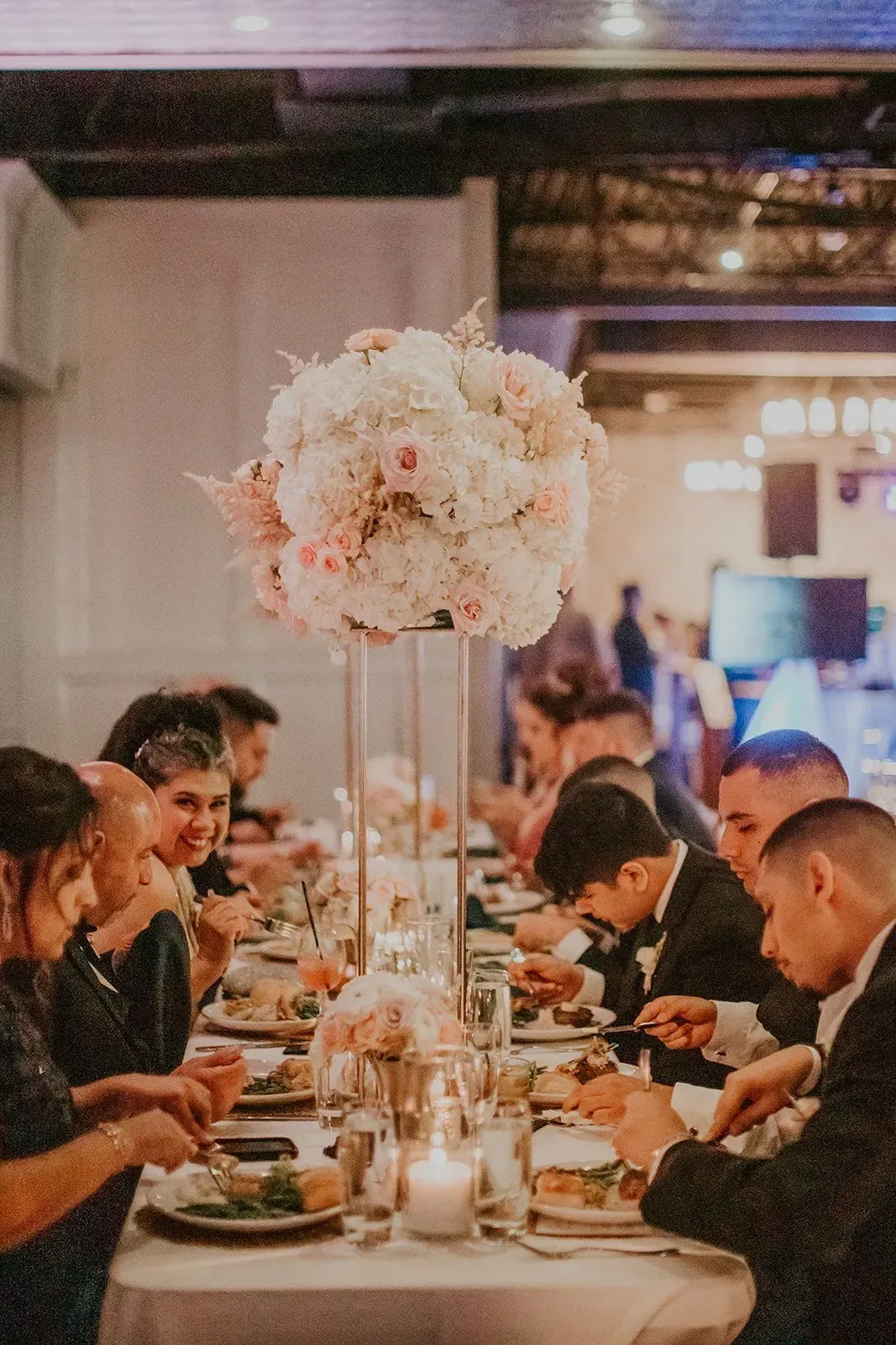 Guests dine at a candlelit banquet table with a tall white floral centerpiece