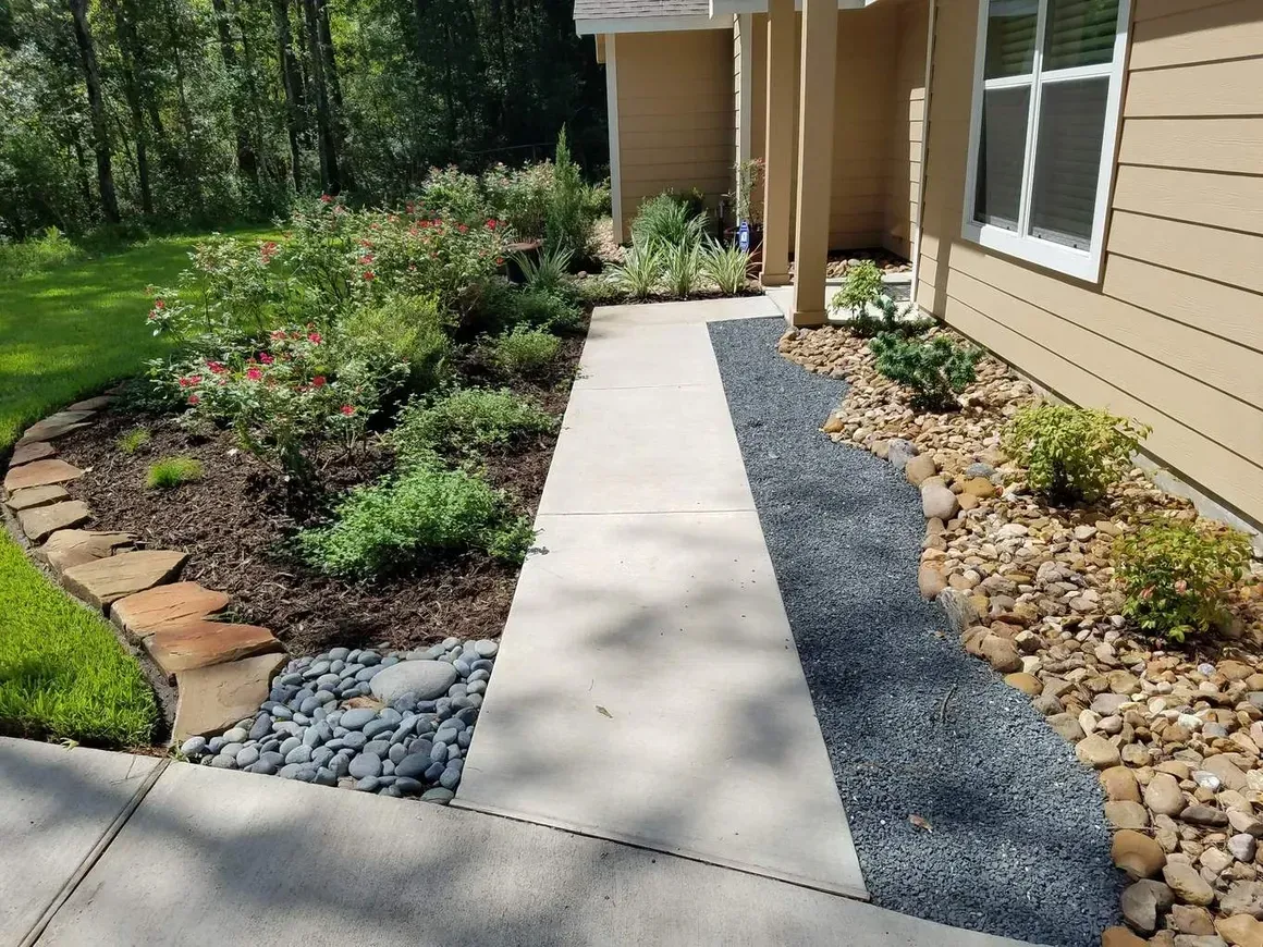Concrete walkway bordered by flower beds and gravel next to a light-colored house.