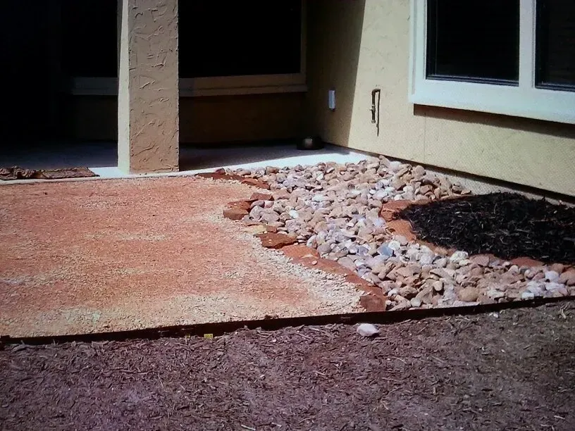 Patio with red gravel, rocks, and brown ground near a house wall.