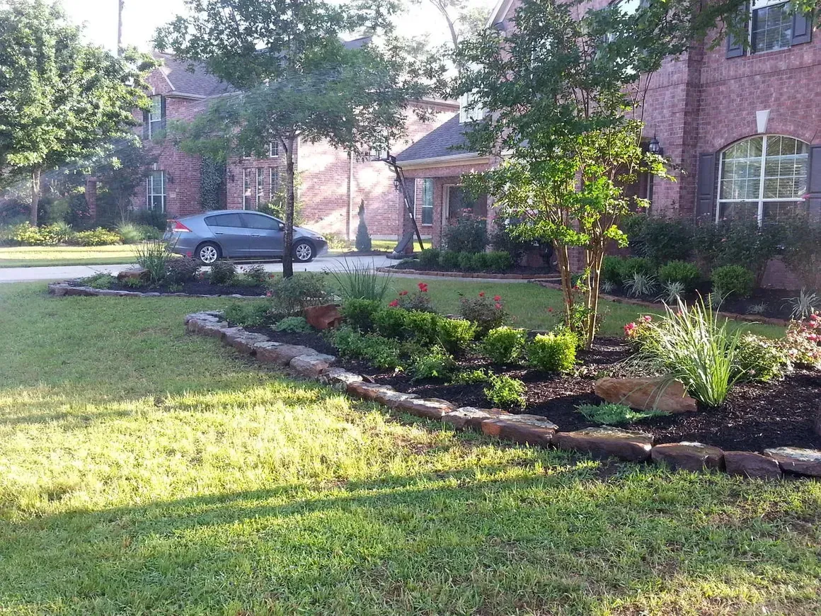 Lush green lawn with a landscaped garden bed in front of a brick house; a car is parked in the driveway.