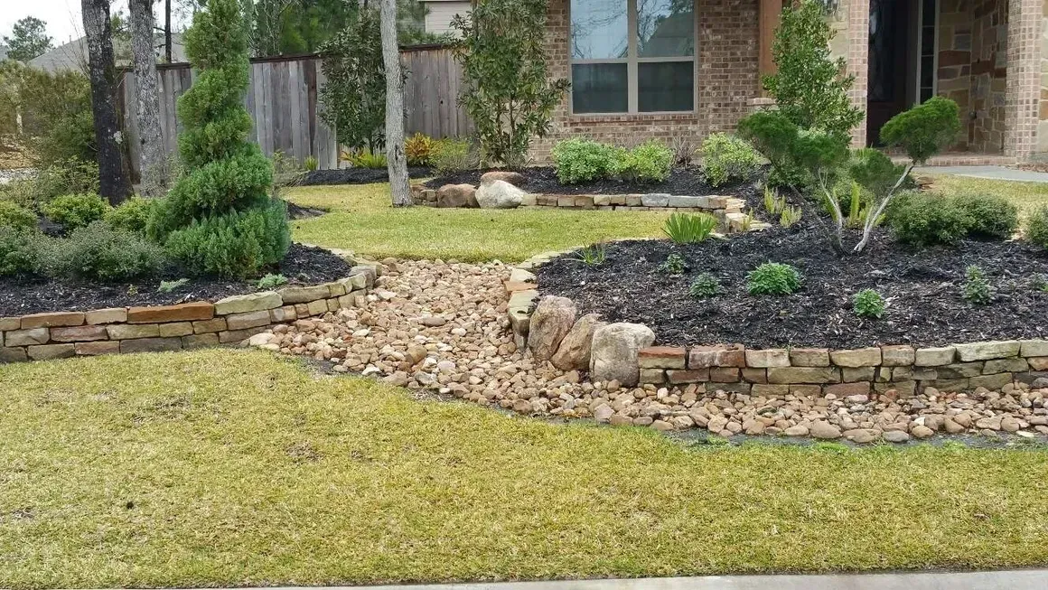 Front yard with a rock-lined dry creek bed, green grass, and brick-edged flower beds with various plants.