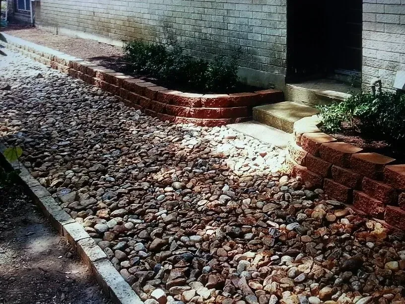 Brick retaining walls frame a rock-covered path leading to a doorway, with planted greenery in the walls.