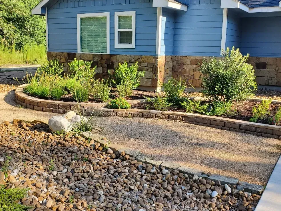 Landscaped pathway and garden bed in front of a blue house with stone foundation.