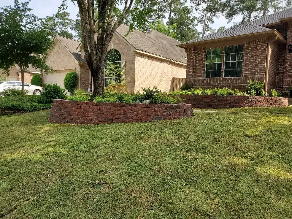 Lawn with a brick retaining wall and trees in front of a brick house with large windows on a sunny day.