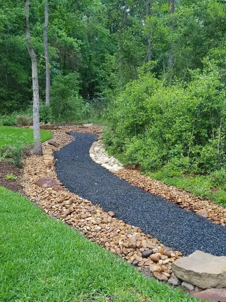 A dark gravel path winds through a garden, bordered by rocks and grass, with trees in the background.