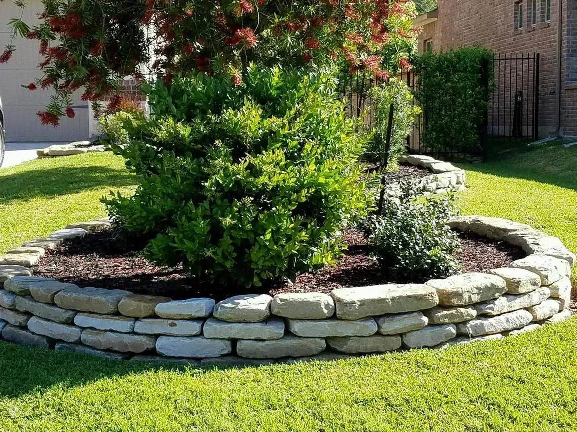 Stone-ringed garden bed with mulch and green plants, set in a grassy yard.