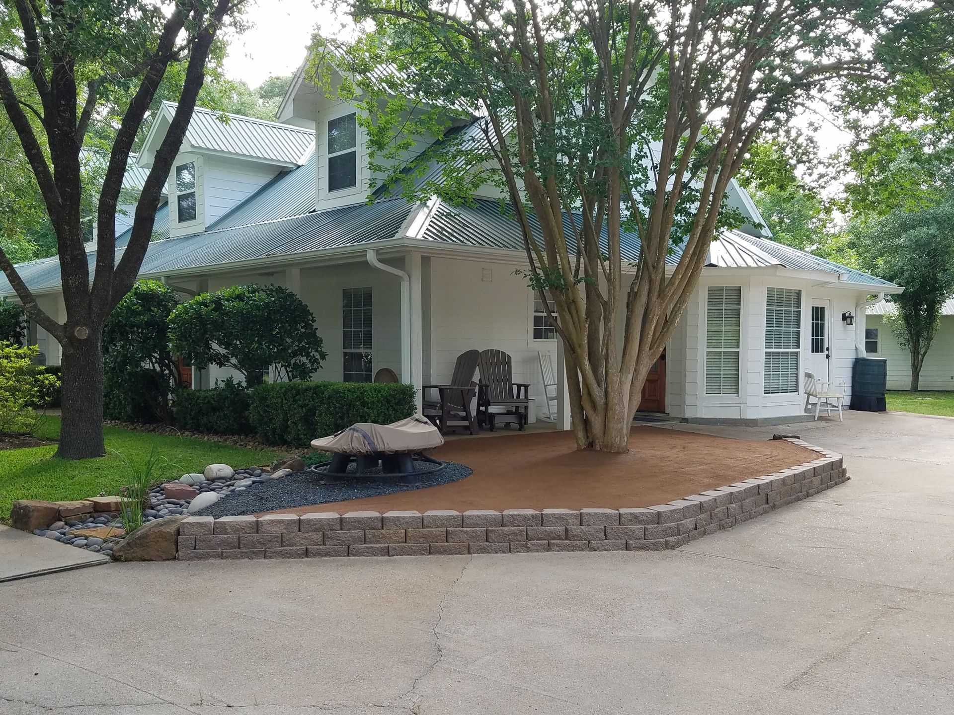 White house with green roof, a landscaped front yard with a rock wall, and a large tree in front.