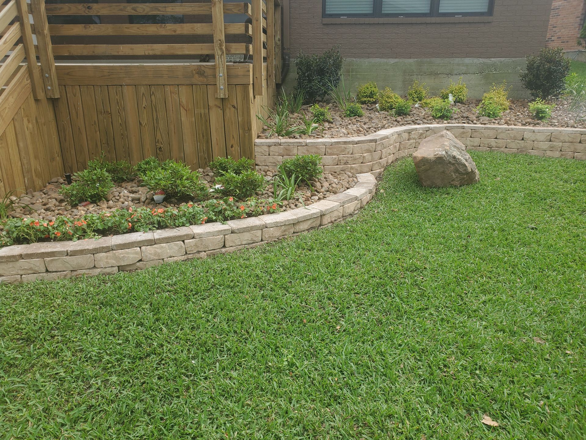 Green lawn bordered by a low retaining wall with flower bed and boulder near wooden structure.