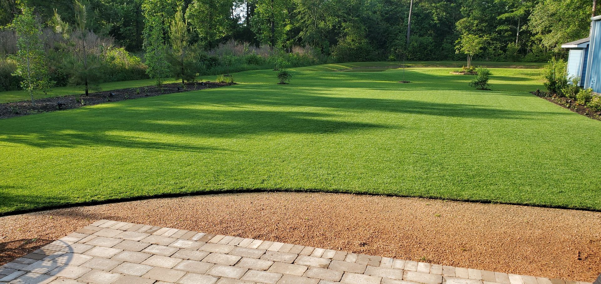 A green lawn bordered by gravel and a stone patio, with trees in the background.