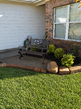 Front porch with black metal bench, brick and siding exterior, and trimmed shrubs by a window.