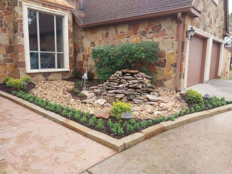 Stone-faced house with landscaped bed containing a rock fountain, gravel, and trimmed bushes, bordered by a concrete curb.
