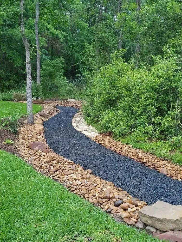 Curving gravel pathway bordered by tan and brown rocks through a green wooded area.