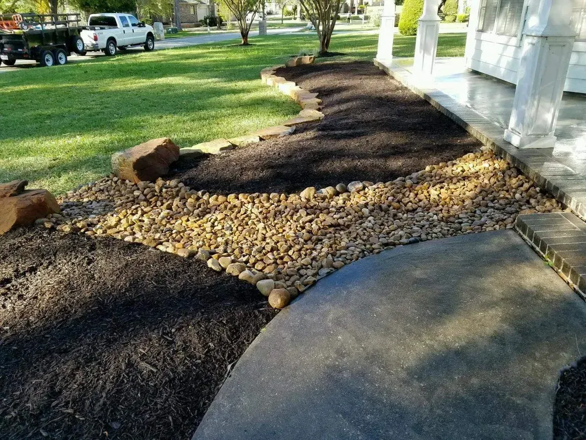 Landscaped front yard with black mulch, tan river rocks, and a concrete path.