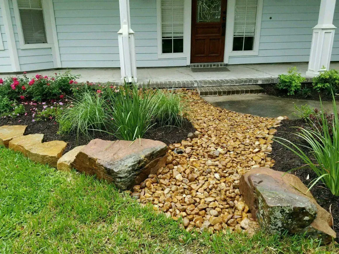 Landscaped front yard with a stone path, rock borders, and plants, leading to a porch and house.