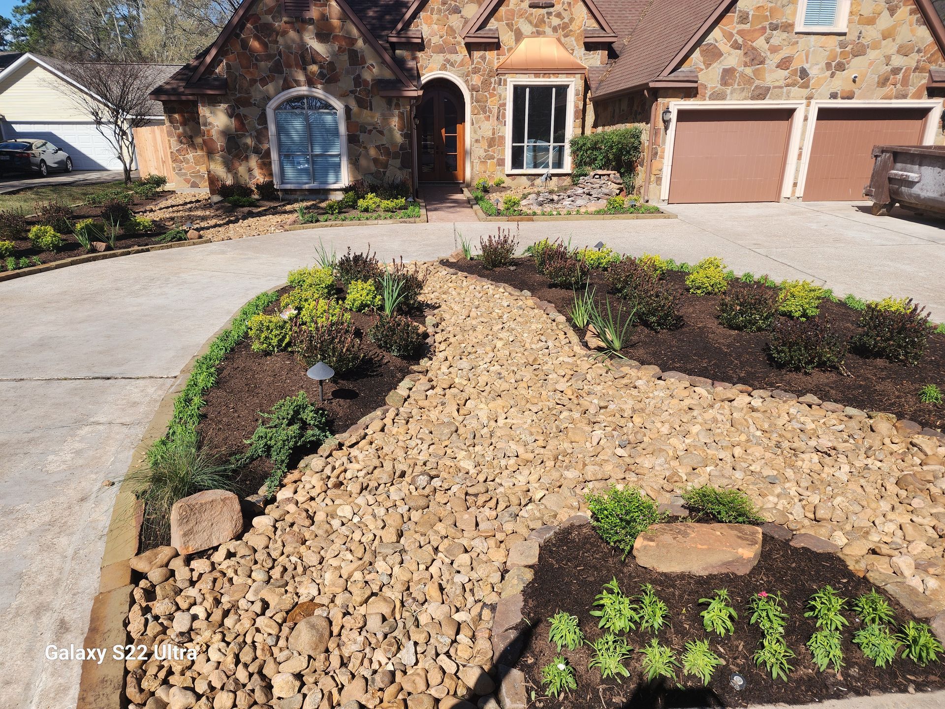 Stone-covered walkway leads to a stone house with brown garage doors and a well-manicured yard with fresh plantings.