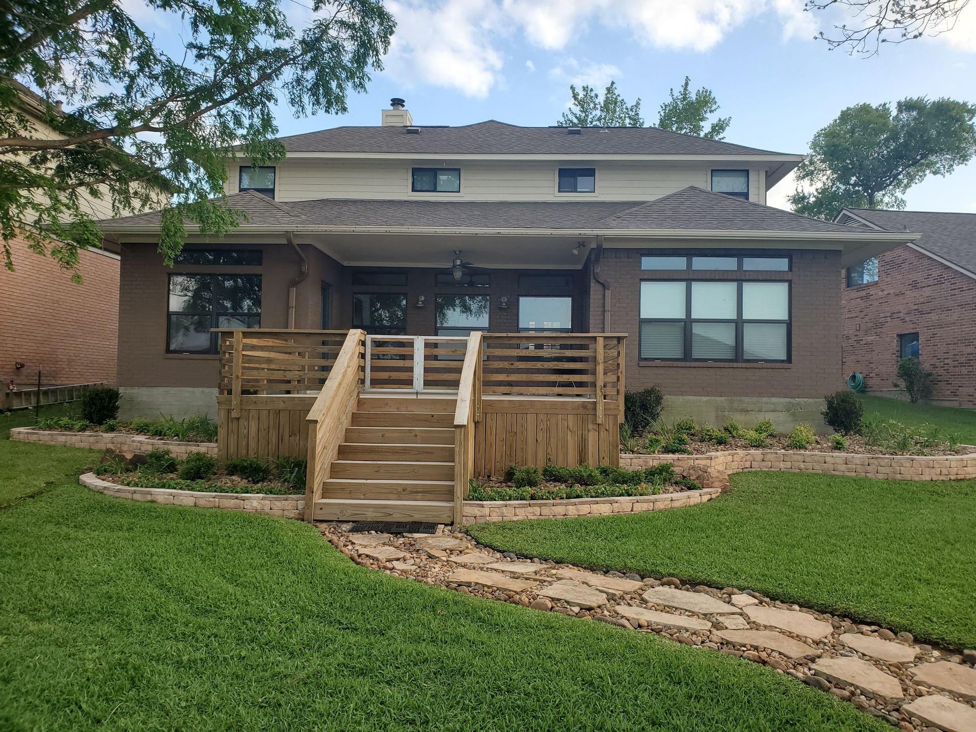 Back of a two-story brick house with a wooden deck, stone path, and green lawn under a blue sky.