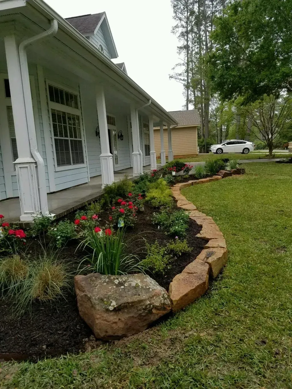 A flower bed with a stone border in front of a white house with a porch.