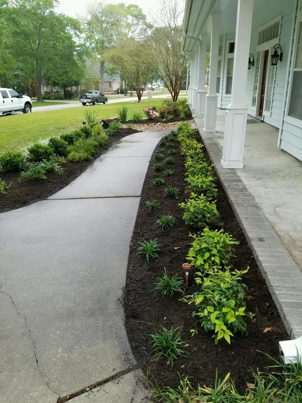 Concrete walkway leading to a house with a porch and flower beds.