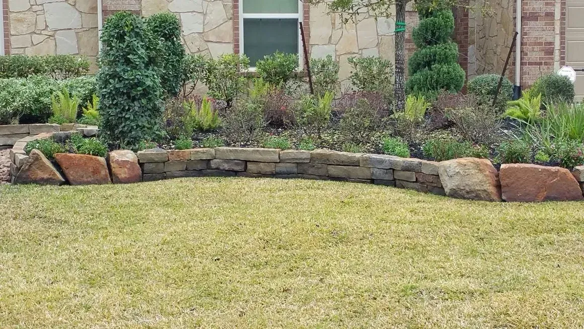 Stone-edged flower bed with various plants and shrubs, in front of a house, on a grassy lawn.