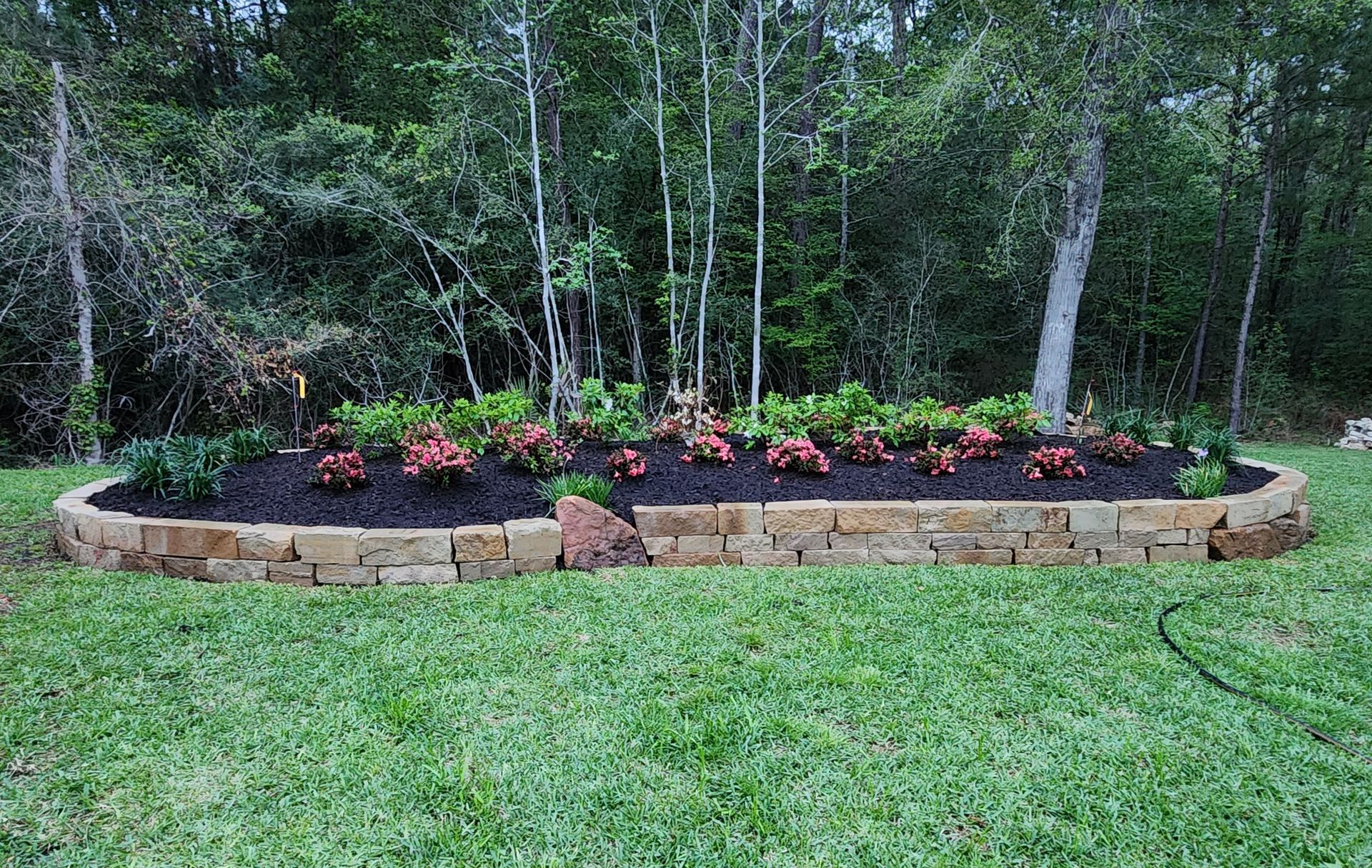 A stone-edged flower bed filled with red and green plants, against a backdrop of trees and a green lawn.