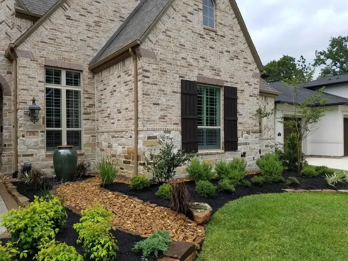 Brick house with landscaped yard, dark brown shutters, green grass, and small plants.