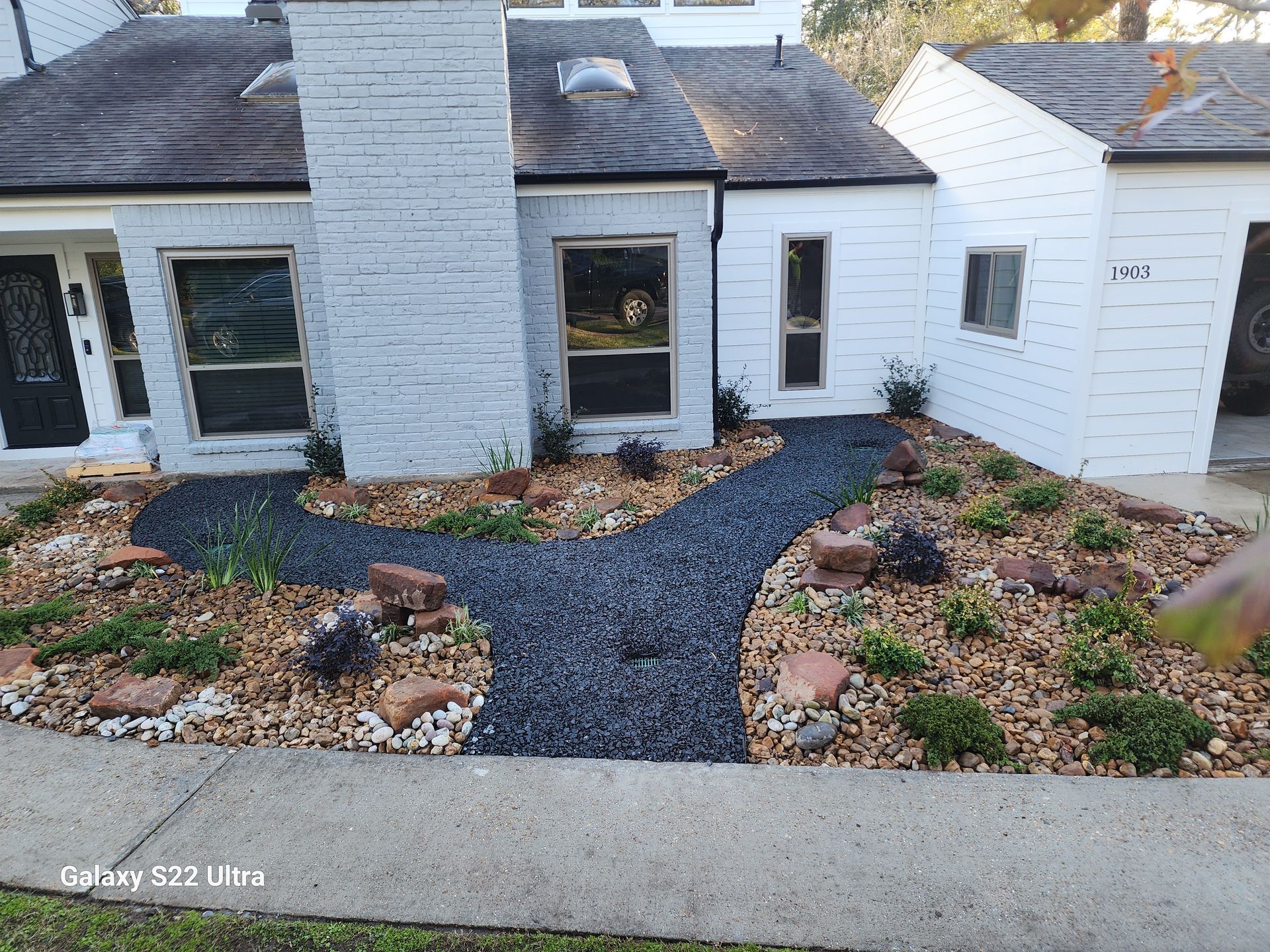Exterior view of a white house with a landscaped yard featuring a black gravel path, rocks, and various plants.
