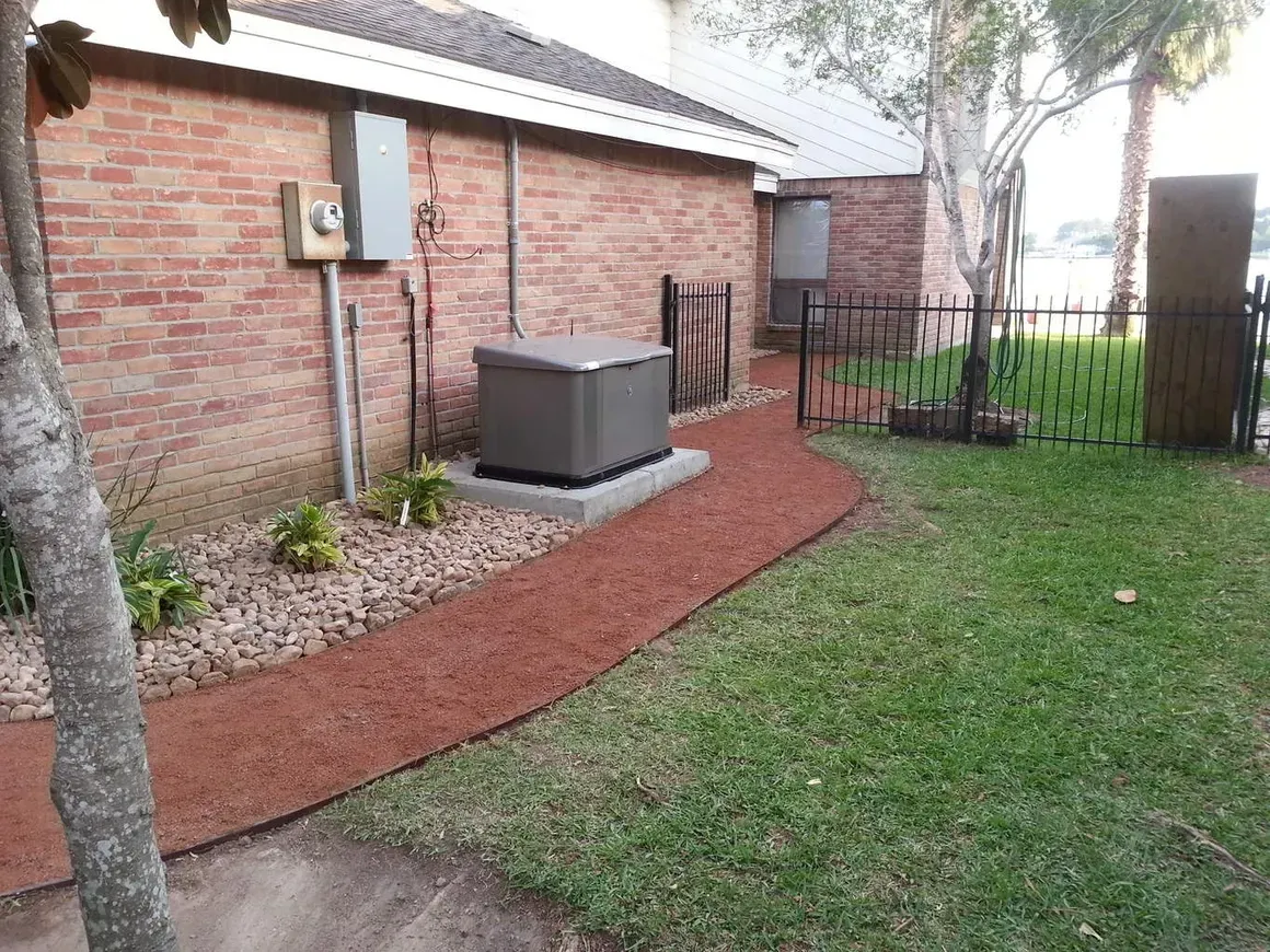 A red gravel pathway next to a brick building, leading to a generator on a concrete pad in a yard.