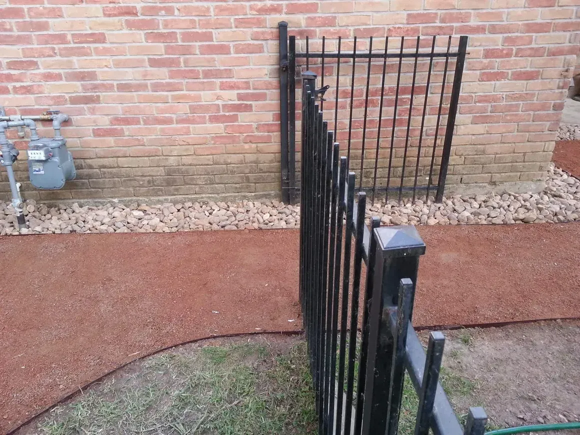 Black metal fence next to a red gravel path against a brick wall. A gas meter is visible.