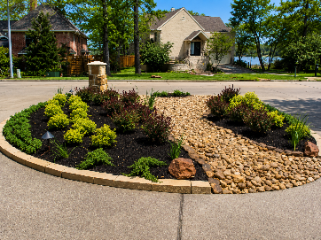 Circular landscaped flower bed with mulch, rocks, and shrubs along a suburban street, with houses in the background