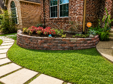 A brick-faced garden bed with a stone walkway leads to a brick house with a green lawn.