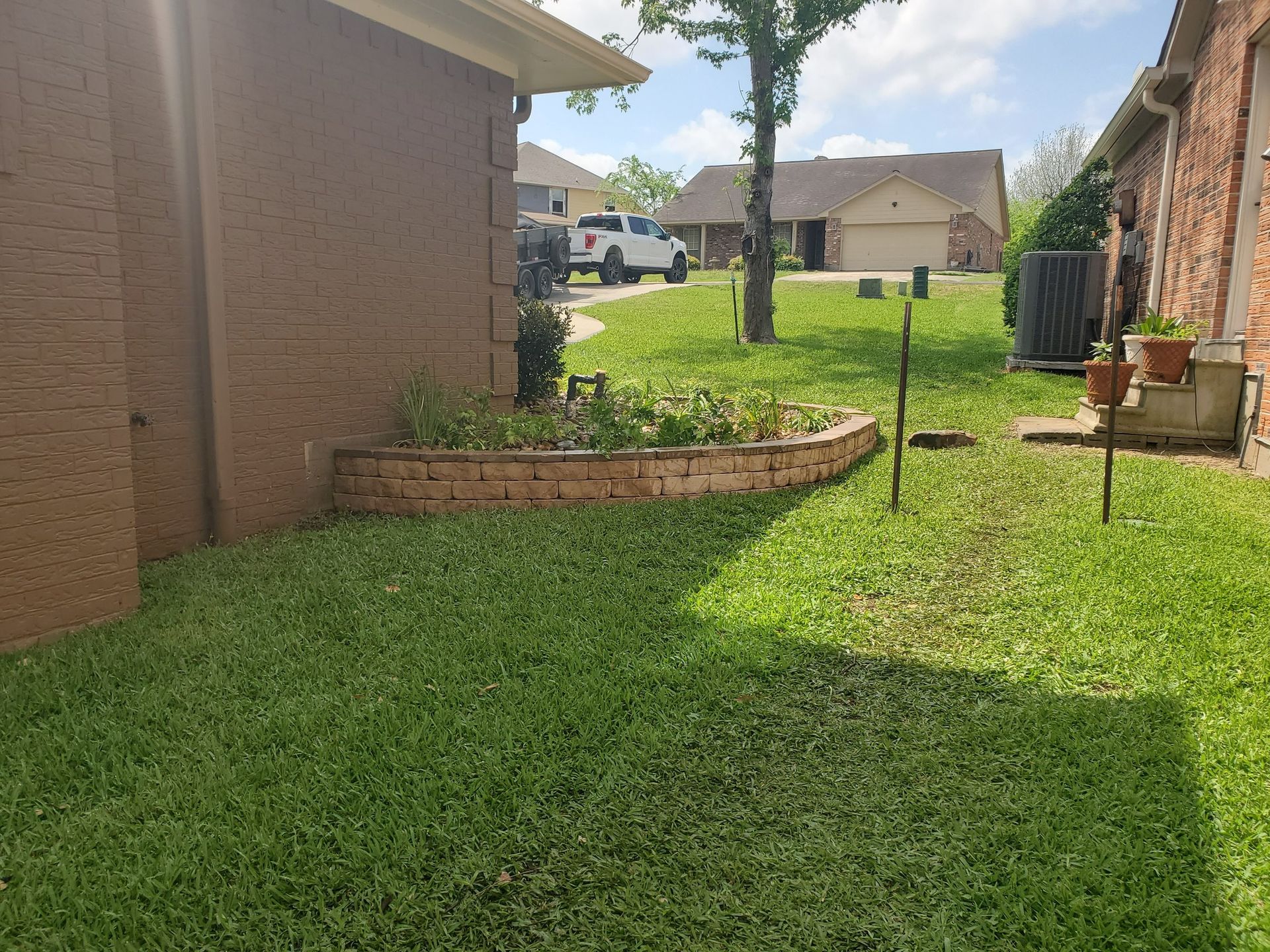 A grassy backyard with a brick planter, brown house, and a pickup truck in the distance.