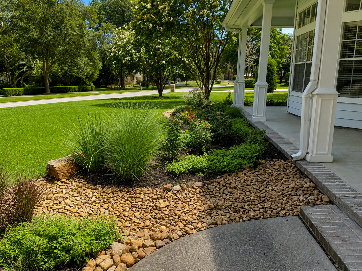 Front porch with white columns beside a landscaped garden of stones, shrubs, and lawn.