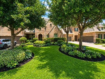 Landscaped brick home with lush green lawn, curved flower beds, and mature trees under bright sun