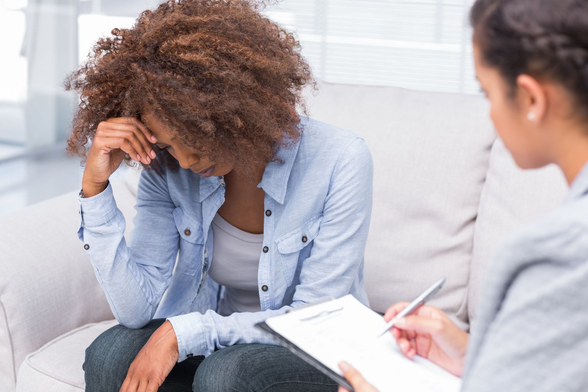 A person sits on a sofa, looking distressed with their head in their hands, while another takes notes on a clipboard.