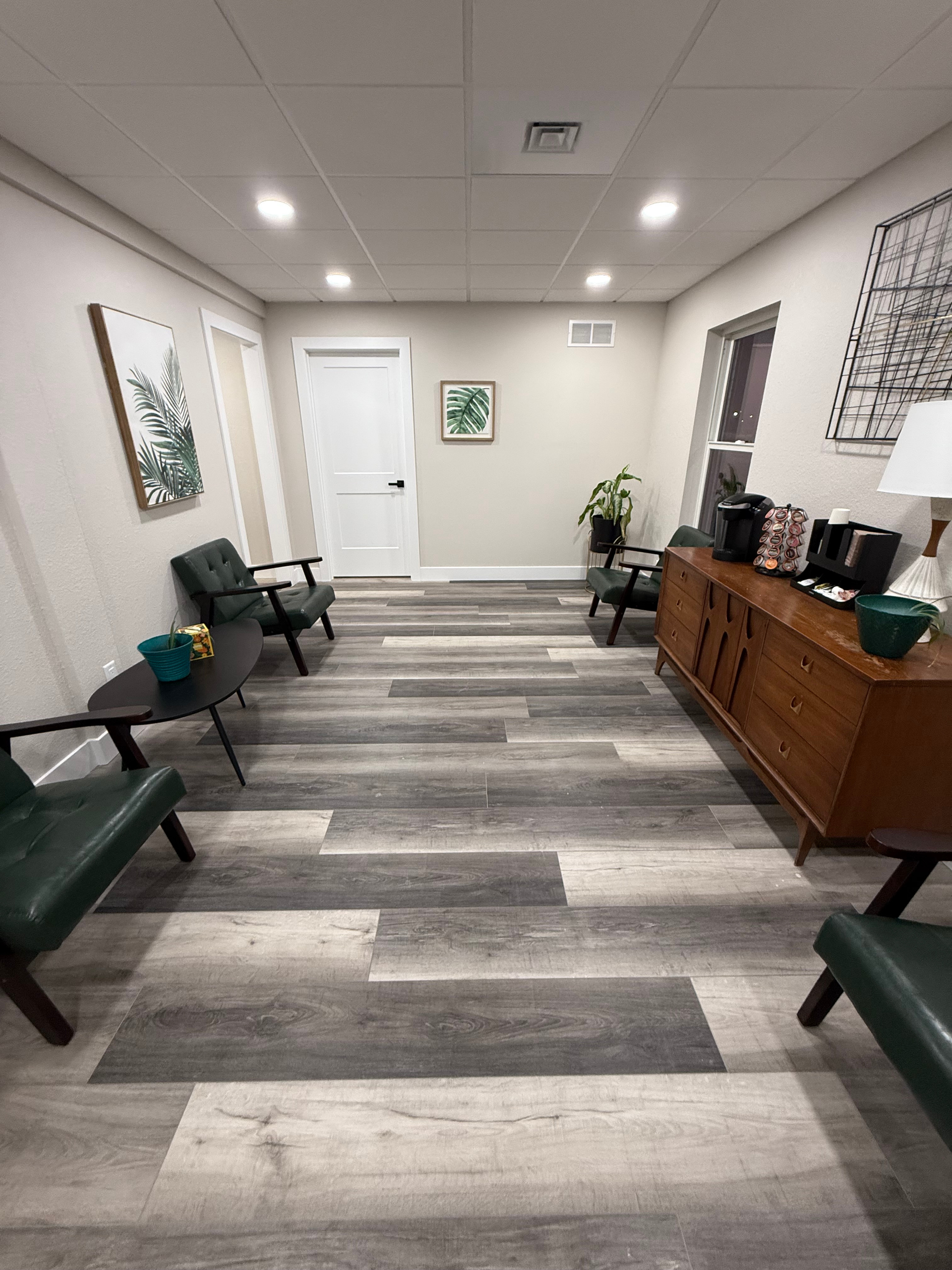 Waiting room with wooden floor, green chairs, and a wooden cabinet with a coffee station.