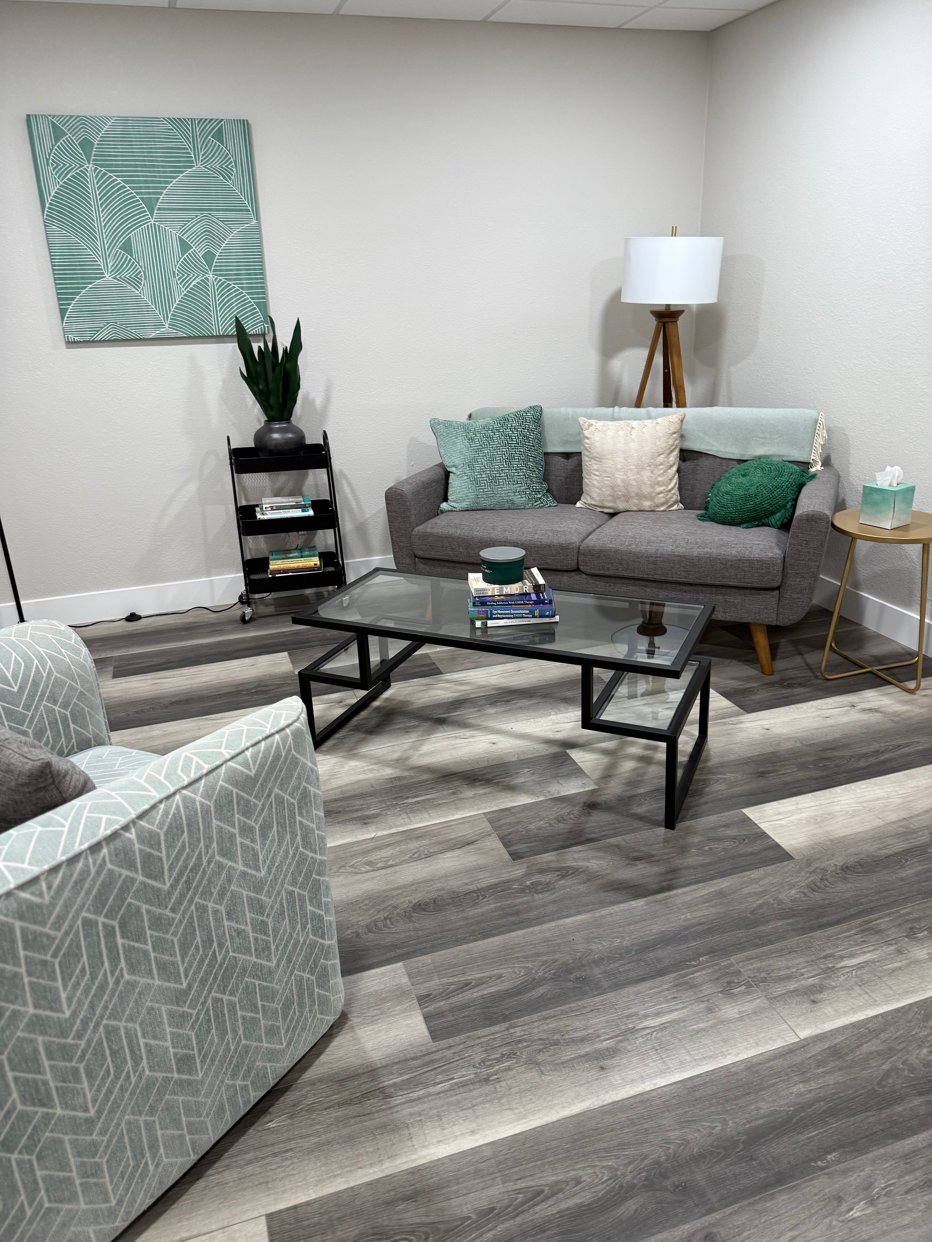 Cozy waiting room with gray furniture, artwork, and a glass coffee table on a wood-look floor.