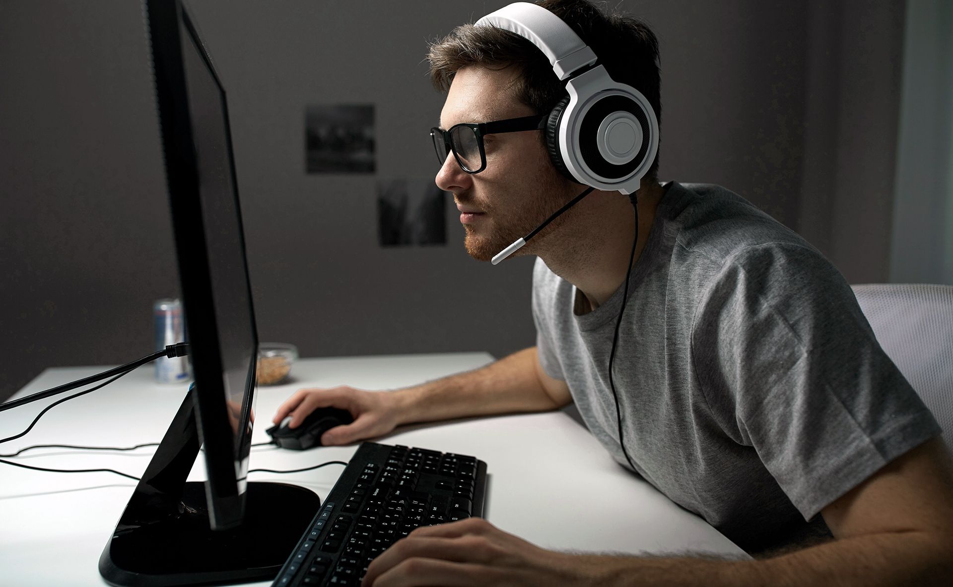 A person wearing glasses and a headset sits in a dimly lit room, using a mouse and keyboard while gaming at a computer.
