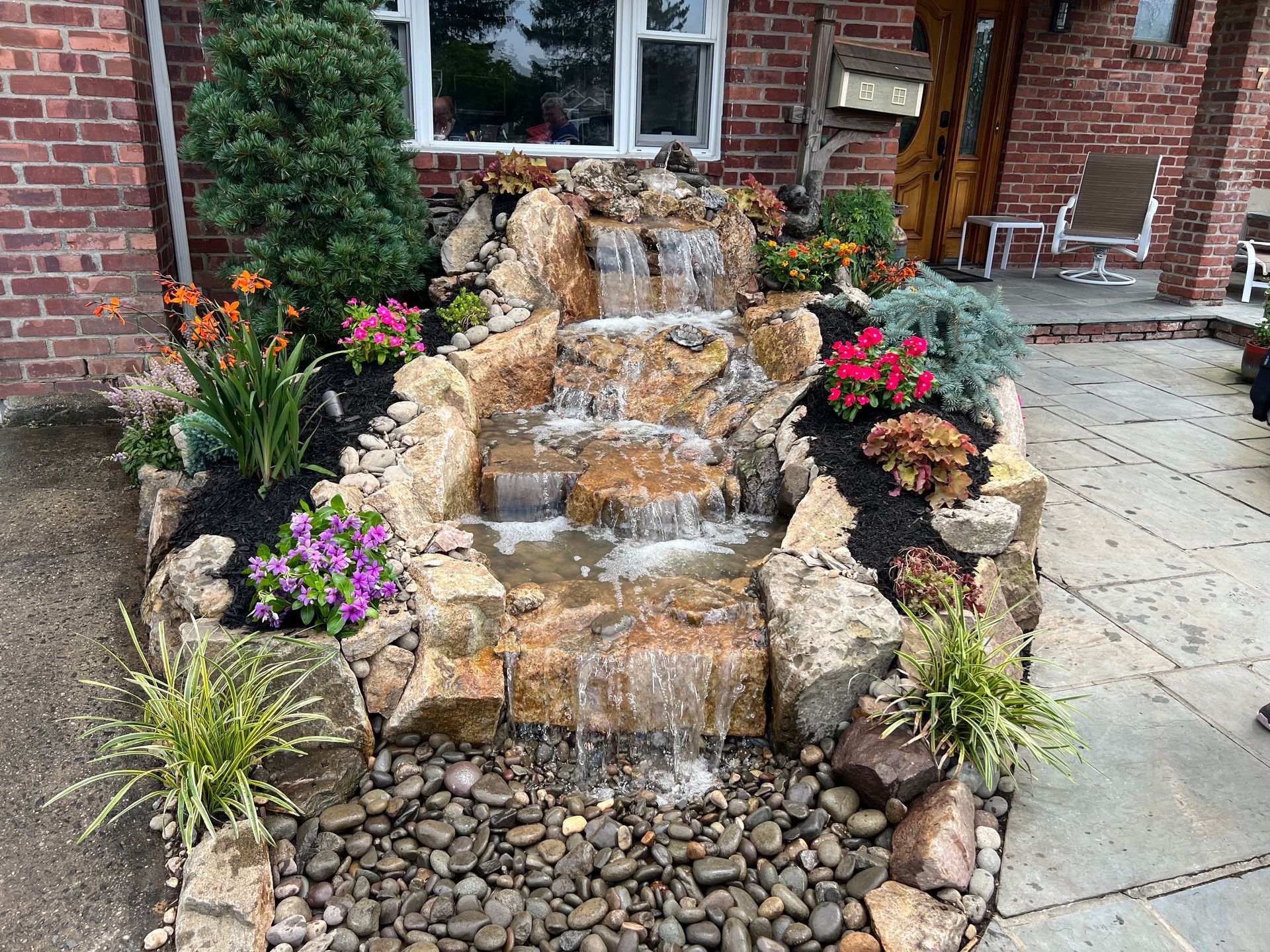 A stone garden waterfall feature with cascading tiers, surrounding colorful flowers and greenery, set on a stone patio.