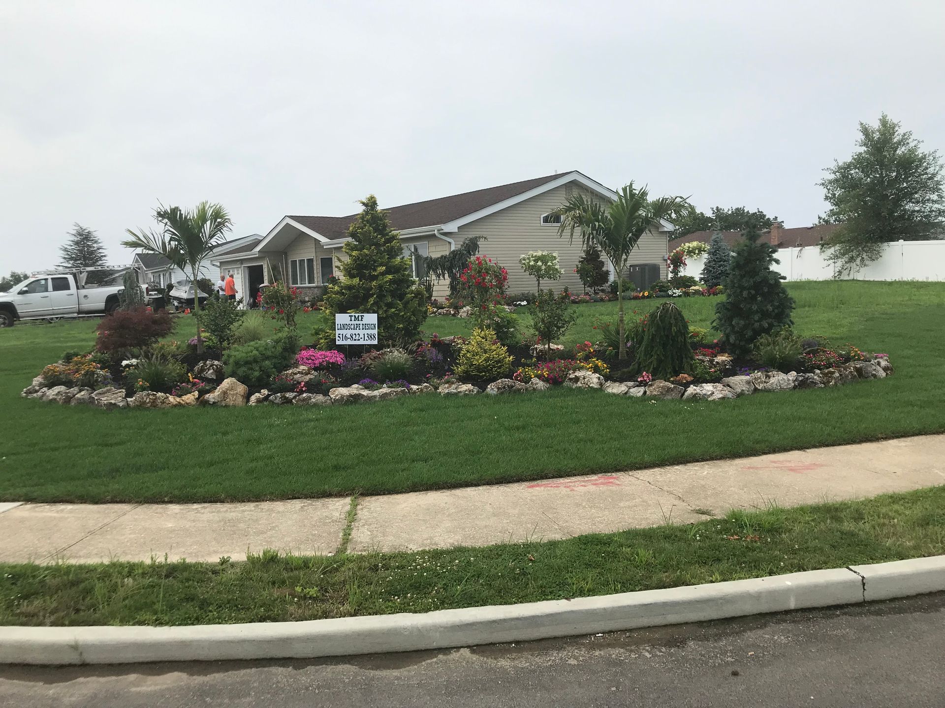 A one-story tan house with a large, landscaped flower bed in the front yard, situated next to a sidewalk and street.
